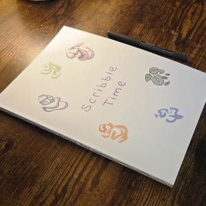 May include: A white notebook with the words "Scribble Time" and colorful illustrations of whimsical shapes. A black pen rests on the top right corner of the notebook, which is placed on a wooden surface.