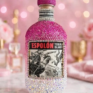 May include: An Espolon Tequila Blanco bottle covered in pink and clear rhinestones in an ombre pattern. The bottle features a central label with a skeleton and rooster illustration, a rhinestone-encrusted cap, and a 750ml volume label.