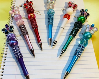 May include: Eight decorative pens with jeweled butterfly accents and colorful beads. The pens are arranged on a lined notebook page.