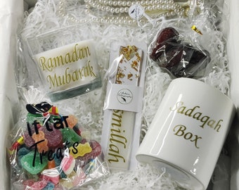 May include: A white gift box filled with Ramadan themed gifts. The box contains a white candle with the text "Ramadan Mubarak", a white mug with the text "Sadaqah Box", a bag of colorful candy with the text "Iftar Treats", a white bookmark with gold accents and the text "Bismillah", and a string of white pearls.