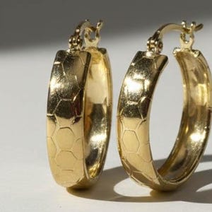 May include: A pair of gold hoop earrings with a honeycomb design. The earrings are made of a shiny gold material. The earrings are displayed on a white surface, casting a shadow.