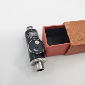 May include: A black and silver camera accessory with the text "LEITZ WETZLAR Germany" and a dial. The accessory is next to a small, open, reddish-brown box. The dial has an arrow pointing to the left.