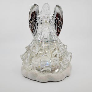 May include: A clear, decorative angel figurine with silver wings. The angel is wearing a tiered dress and sits on a glittery, cloud-shaped base. The wings have a reflective, metallic finish. The figurine is designed for decorative purposes.