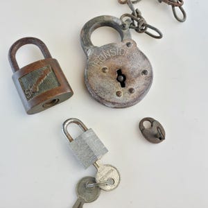 May include: Four vintage padlocks with keys. One padlock is silver and has a keyhole. Another padlock is brass and has a keyhole. The third padlock is brass and has a keyhole. The fourth padlock is brass and has a keyhole. The padlocks are on a white surface.