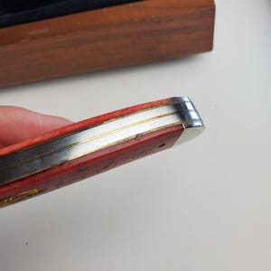 Puede incluir: Primer plano de una navaja de bolsillo roja y plateada. El cuchillo tiene un mango rojo con detalles plateados y dorados. La hoja est&aacute; cerrada y el extremo del cuchillo es plateado.