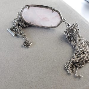 May include: A silver chain bracelet with a pink stone pendant and a tassel charm. The stone is a light pink color and has a smooth, polished finish. The tassel charm is made of silver chains and is about 3 inches long.