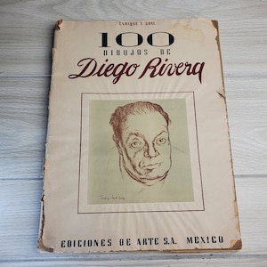 May include: A vintage book cover with the title "100 Dibujos de Diego Rivera" in red lettering. The cover features a black and white drawing of Diego Rivera, a famous Mexican artist.