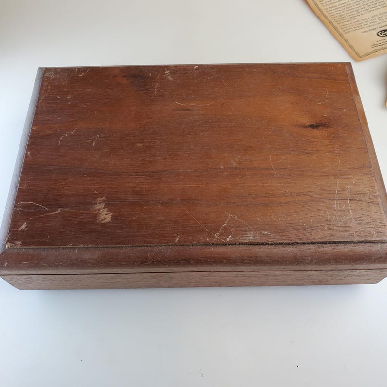 Puede incluir: Una caja rectangular de madera con un acabado marr&oacute;n oscuro. La caja muestra signos de desgaste, con ara&ntilde;azos y marcas en la superficie. Los bordes est&aacute;n bien definidos y el dise&ntilde;o general es simple y cl&aacute;sico. La caja parece ser de madera maciza.