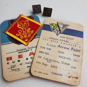 May include: Vintage Cub Scout memorabilia, including a red and yellow patch with a bear design and text, two small metal pins, and two aged cards. One card reads "Silver Arrow Point" and "Galveston, Texas."