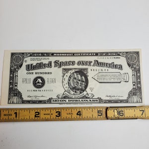May include: A black and white commemorative currency certificate with the text "Unified Space over America" and "Apollo Eleven" with a picture of the Apollo 11 crew. The certificate is a "Moondust Certificate" and has a serial number of "NRG 2 XLR 8".