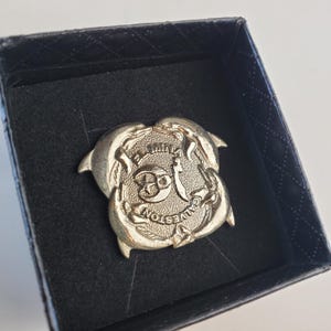 May include: A gold-colored metal pin with two dolphins intertwined around a circle with the text "Galveston Island", "The", and "Smile" inside. The pin is in a black velvet box.