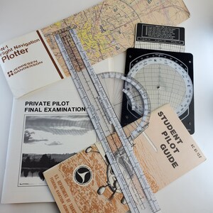 May include: A collection of aviation tools and guides for pilots, including a flight navigation plotter, a protractor, a student pilot guide, and a map. The tools are laid out on a white surface.
