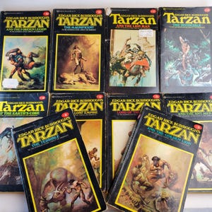 May include: A collection of vintage paperback books featuring the character Tarzan. The covers display illustrations of Tarzan in various action poses, with titles like "The Magnificent" and "The Golden Lion". The books have a black background with yellow and white text.