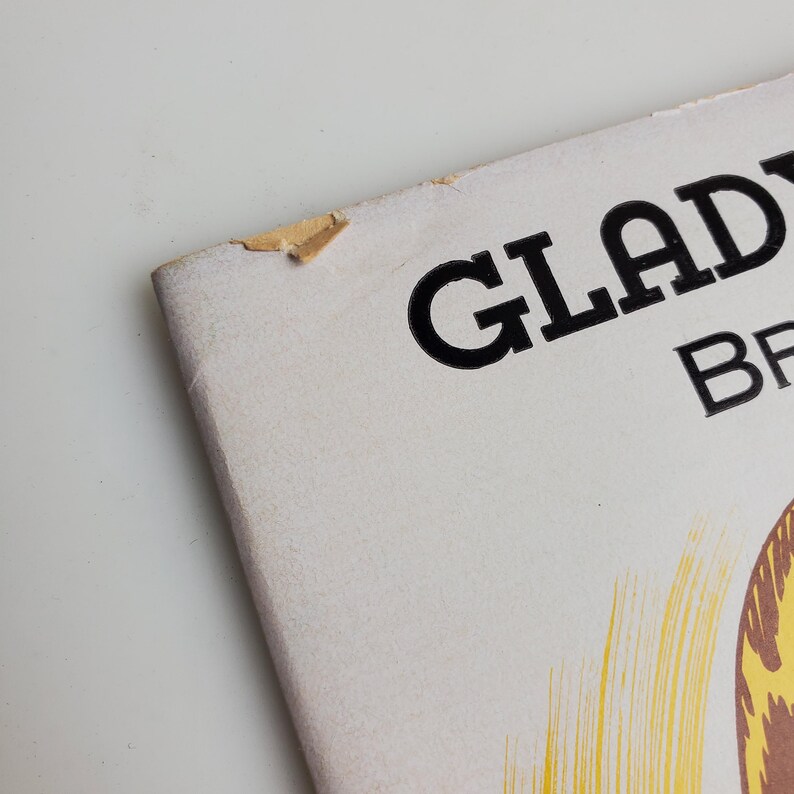 Op de afbeelding: Een vintage boekomslag met de titel "GLADYS BR" in zwarte tekst op een witte achtergrond. De omslag heeft een gele en bruine illustratie van haar.