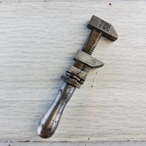 Puede incluir: Una llave inglesa ajustable de plata vintage con mango negro. La llave tiene el texto "GEM T.M.L." en la parte superior.