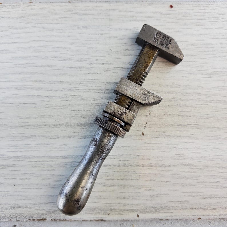 Puede incluir: Una llave inglesa ajustable de plata vintage con mango negro. La llave tiene el texto "GEM T.M.L." en la parte superior.