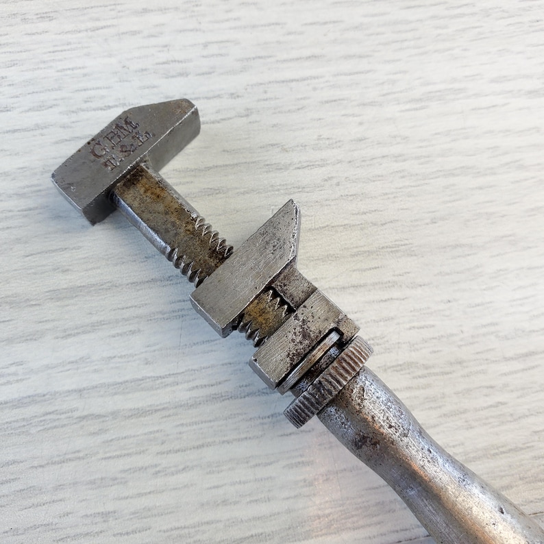 Puede incluir: Una llave inglesa ajustable vintage con acabado plateado y el texto "GEM MFG. & CO." en el mango.