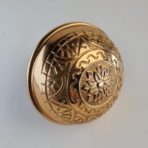 May include: A gold-colored metal doorknob with an intricate, embossed design. The doorknob has a circular shape with a raised, floral pattern in the center.