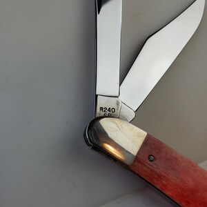 Puede incluir: Una navaja de bolsillo parcialmente abierta con una hoja de acero pulido y un mango de madera marr&oacute;n rojizo. El cuchillo tiene el texto "R240" en el metal. La hoja est&aacute; parcialmente abierta, revelando el filo. El mango es de color marr&oacute;n rojizo c&aacute;lido.