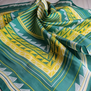 May include: A green and yellow patterned silk scarf with a geometric design. The scarf is folded and draped on a light wood surface.