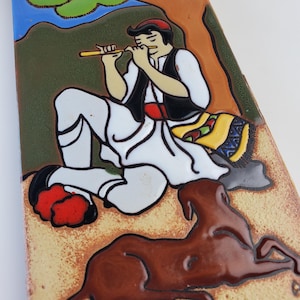 May include: A ceramic tile featuring a seated figure playing a flute, dressed in traditional white pants, a black vest, and a red cap. A brown dog rests below. The background includes green, brown, and blue tones.