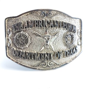 May include: Silver-toned metal belt buckle with the text "THE AMERICAN LEGION DEPARTMENT OF TEXAS." The buckle features a star with a longhorn skull, and two circular emblems. The buckle has a textured background and decorative scrollwork.