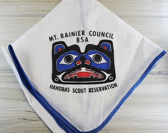 Pode incluir: Um lenço branco com uma borda azul. Apresenta um rosto de animal estilizado em preto e azul com acentos vermelhos. O texto "MT. RAINIER COUNCIL BSA" e "HAHOBAS SCOUT RESERVATION" está impresso no lenço.