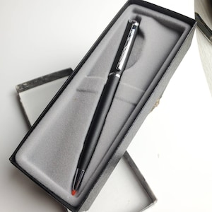 Puede incluir: Un bolígrafo negro con detalles plateados y punta roja, presentado en una caja negra con interior gris. El bolígrafo tiene un diseño elegante, adecuado para uso profesional o como regalo.
