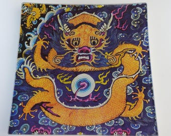 Puede incluir: Plato decorativo cuadrado con una detallada ilustración de un dragón dorado sobre un fondo azul oscuro. El dragón tiene rasgos intrincados y está rodeado de nubes y patrones arremolinados. El plato tiene un acabado transparente y brillante.