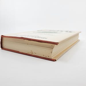 May include: A hardback book with a beige cover and a red spine. The title and author's name are printed on the cover. The book's pages are visible, and the edges show some wear. The book is lying on a white surface.