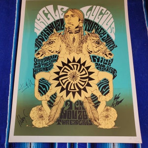 May include: A vintage concert poster with a turquoise and green background featuring a Native American figure with a headdress and a large yellow sunburst. The poster advertises a concert featuring Iggy Pop,  Strayhan and the Good Neighbors,  Nick and Cage, and 2 Chuz  Threadgills. The poster is signed by Iggy Pop, Strayhan, and Nick.