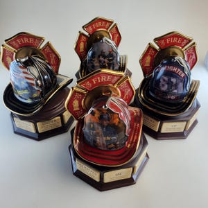 May include: Set of four porcelain firefighter helmet figurines. Each helmet is decorated with a different design, including a firefighter in action, an American flag, and a firefighter's badge. The bases of the figurines are decorated with gold accents and inscriptions.