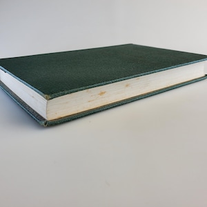 May include: A closed, hardcover book with a dark teal cover. The book's pages are off-white, and the edges show slight discoloration. The cover has a textured appearance, and the book is lying flat on a white surface.