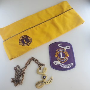 May include: A yellow fabric hat with a Lions Club emblem. A purple patch with the Lions Club emblem and the words "Lions Club International Lioness Club" is also shown. A gold chain necklace with a gold pendant that says "Lioness Club" is also shown.