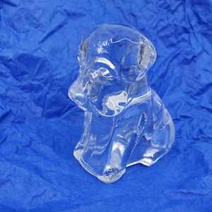 May include: A clear glass figurine of a dog in a sitting position. The sculpture has a smooth, polished surface and detailed features, including the dog's ears, snout, and paws. The figurine is set against a blue background.