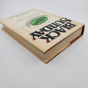 May include: A hardback book titled "Black Sunday" by Thomas Harris. The cover displays a football stadium graphic in green and white, with the title in large black letters outlined in red. The book has a tan cover and brown edges.