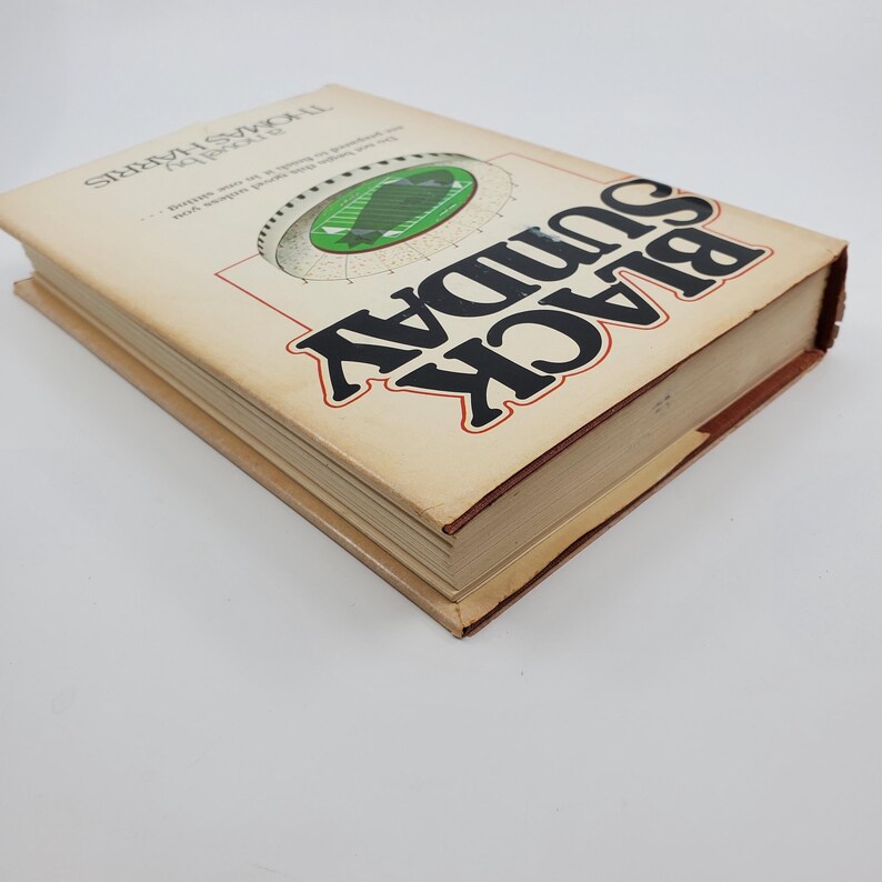 May include: A hardback book titled "Black Sunday" by Thomas Harris. The cover displays a football stadium graphic in green and white, with the title in large black letters outlined in red. The book has a tan cover and brown edges.