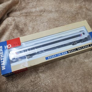May include: A Walthers HO scale model railroad passenger car in a rectangular cardboard box. The packaging features blue, red, and white branding with the text Walthers, HO Scale, and Ready-To-Run Model Railroad Car. The car is white with a dark stripe.