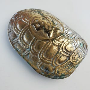 May include: A vintage, oval-shaped brass object with an embossed design. The brass has a tarnished, aged appearance with hints of green oxidation. The design features a central figure with surrounding floral or leaf-like patterns.