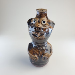 May include: A handmade ceramic bottle shaped like a smiling bear. The brown and tan glazed bottle has a detailed face with blue eyes, ears, and a buttoned shirt. The bottle is a unique piece of art.