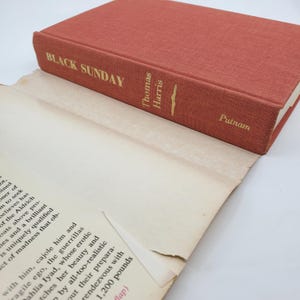 May include: A hardback book titled "Black Sunday" by Thomas Harris, published by Putnam. The book has a reddish-orange cover with gold lettering. The pages are off-white and slightly aged, with text visible.