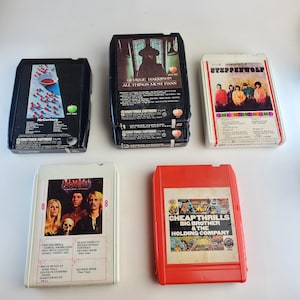 May include: A collection of five vintage 8-track tapes. The tapes feature album art from various artists, including George Harrison, Steppenwolf, and Black Sabbath. The tapes are in various colors, including black, white, and red.