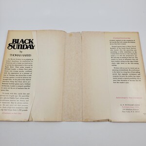May include: An open book titled "Black Sunday" by Thomas Harris. The cover displays the title in bold black font. The pages are off-white, with text detailing the plot and author's biography.