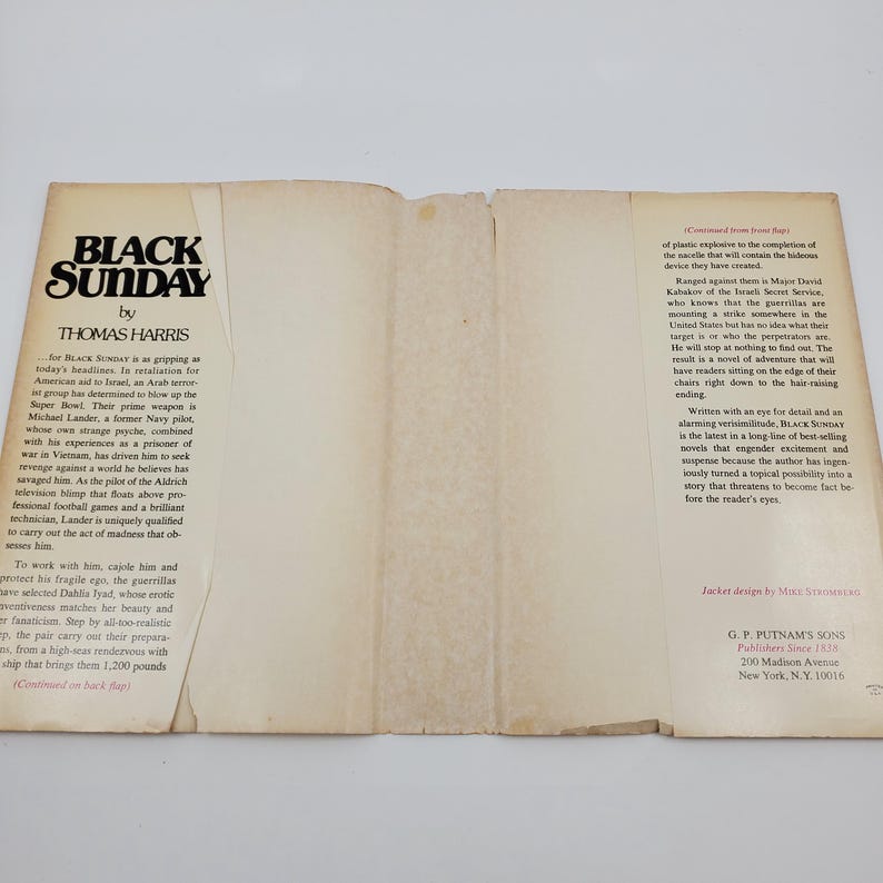 May include: An open book titled "Black Sunday" by Thomas Harris. The cover displays the title in bold black font. The pages are off-white, with text detailing the plot and author's biography.