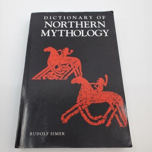 May include: A hardcover book titled "Dictionary of Northern Mythology" by Rudolf Simek. The cover is black with red illustrations of figures on horseback. The book is closed and lying flat.