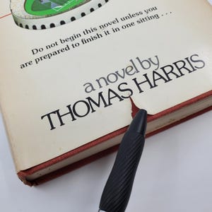 May include: A cream-coloured hardcover book with the title "a novel by THOMAS HARRIS" printed in black. The book's edges are red. The text "Do not begin this novel unless you are prepared to finish it in one sitting..." is printed at the top.