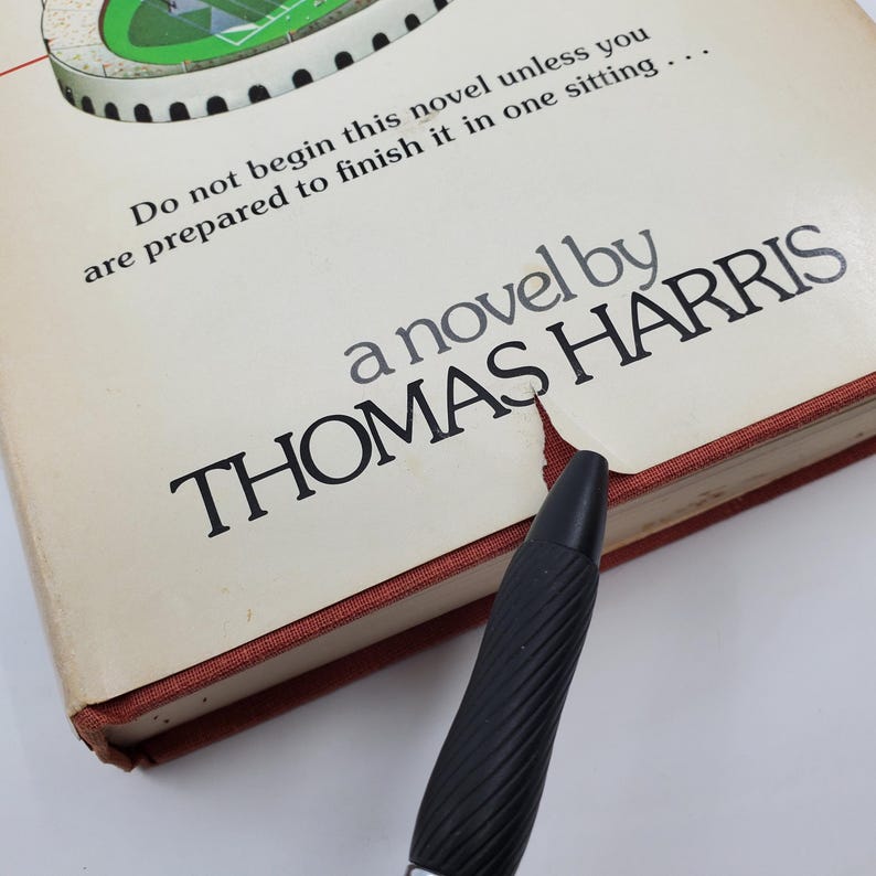 May include: A cream-coloured hardcover book with the title "a novel by THOMAS HARRIS" printed in black. The book's edges are red. The text "Do not begin this novel unless you are prepared to finish it in one sitting..." is printed at the top.