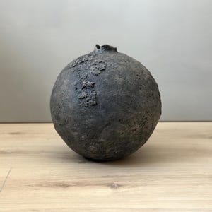 May include: A dark grey, spherical ceramic vase with a textured surface. The vase has a rough, organic appearance, with raised areas and a small, irregular opening at the top. The vase sits on a light-coloured wooden surface.