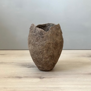 May include: A brown, egg-shaped vase with a textured, rough surface and a jagged top edge. The vase is set against a grey background and rests on a light-coloured wooden surface. The vase is likely made of a natural material.