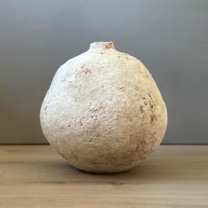 May include: A spherical vase with a narrow opening at the top, featuring a textured, off-white surface with visible earthy brown undertones and a rough, matte finish that gives the object a weathered, organic appearance throughout its body.